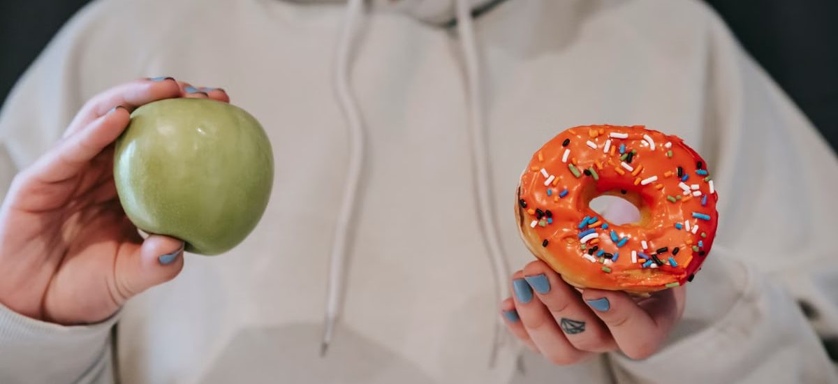 Crop unrecognizable female in loose hoodie showing healthy green apple and sweet delicious donut