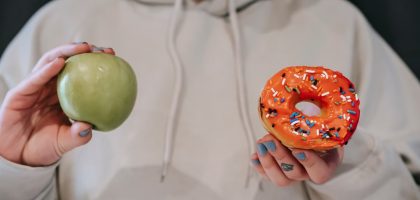 Crop unrecognizable female in loose hoodie showing healthy green apple and sweet delicious donut