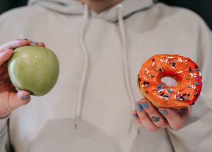 Crop unrecognizable female in loose hoodie showing healthy green apple and sweet delicious donut